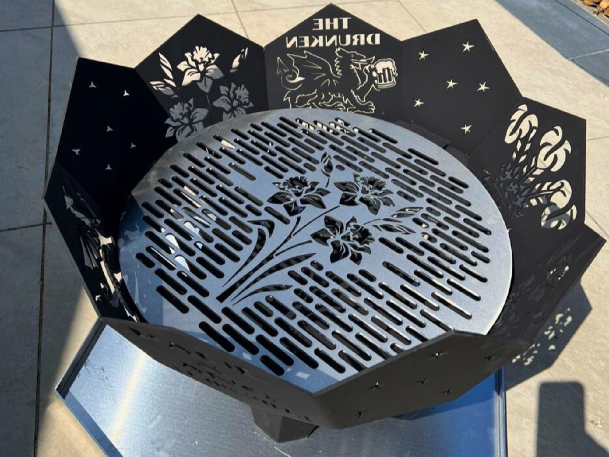 Decorative laser‑cut steel fire pit with floral patterns and griffin artwork casting shadows on outdoor tiles