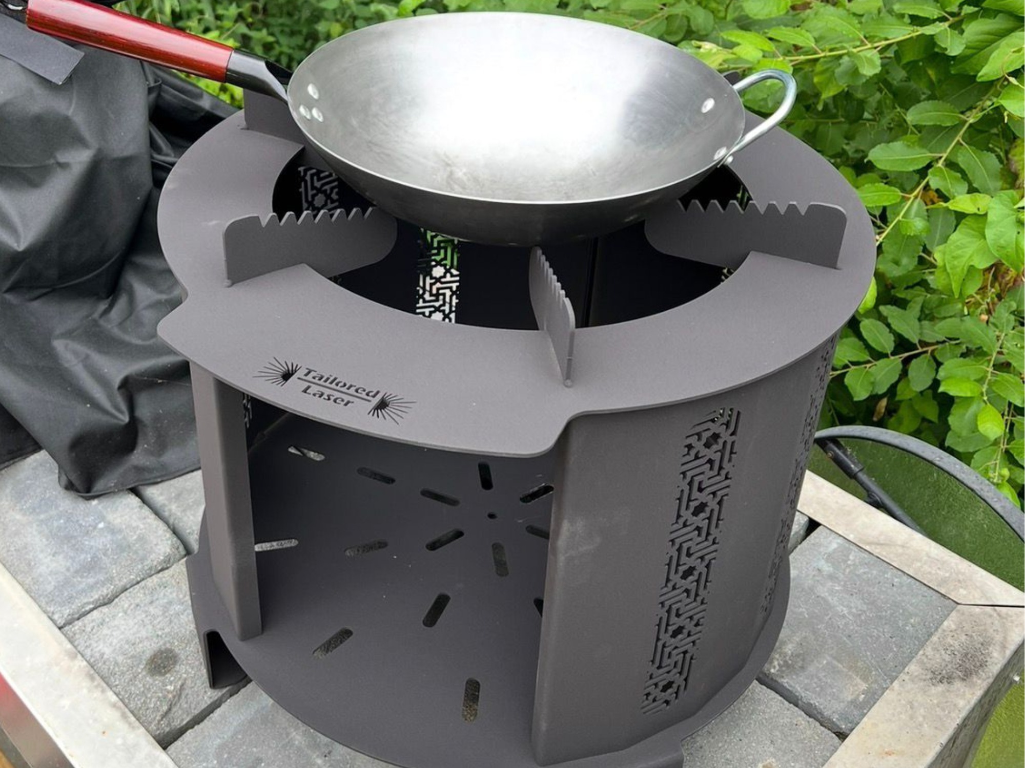 Laser‑cut steel outdoor cooking stove with wok on top, featuring Tailored Laser branding and decorative cutouts