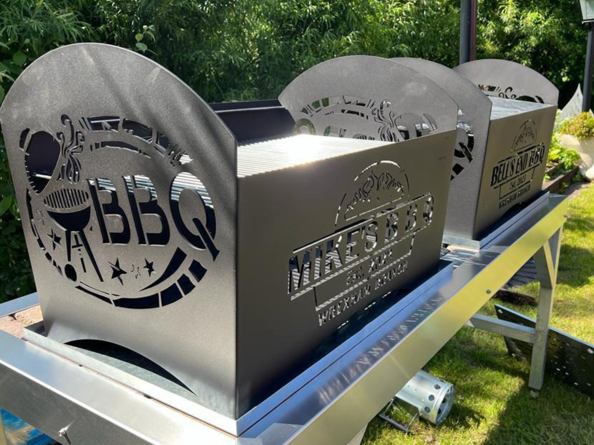 Custom laser‑cut steel BBQ grills with personalised names and decorative cut outs displayed outdoors.