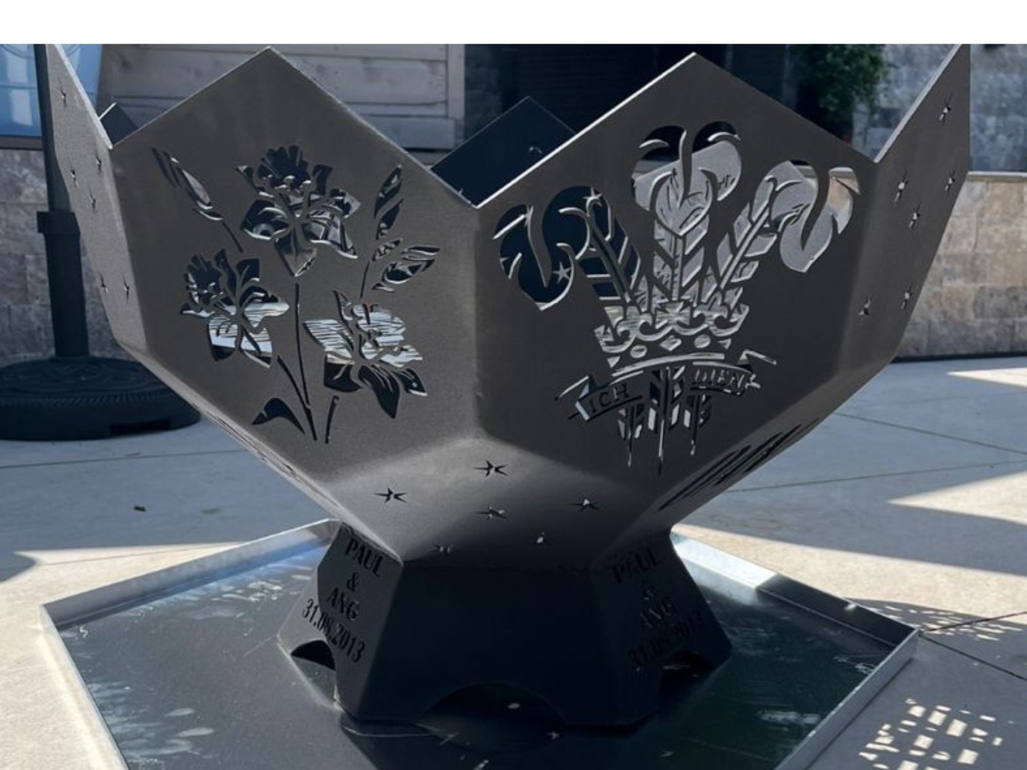 Hexagonal laser‑cut steel fire pit with floral panels, ICH DIEN crest, and personalised engraved base outdoors