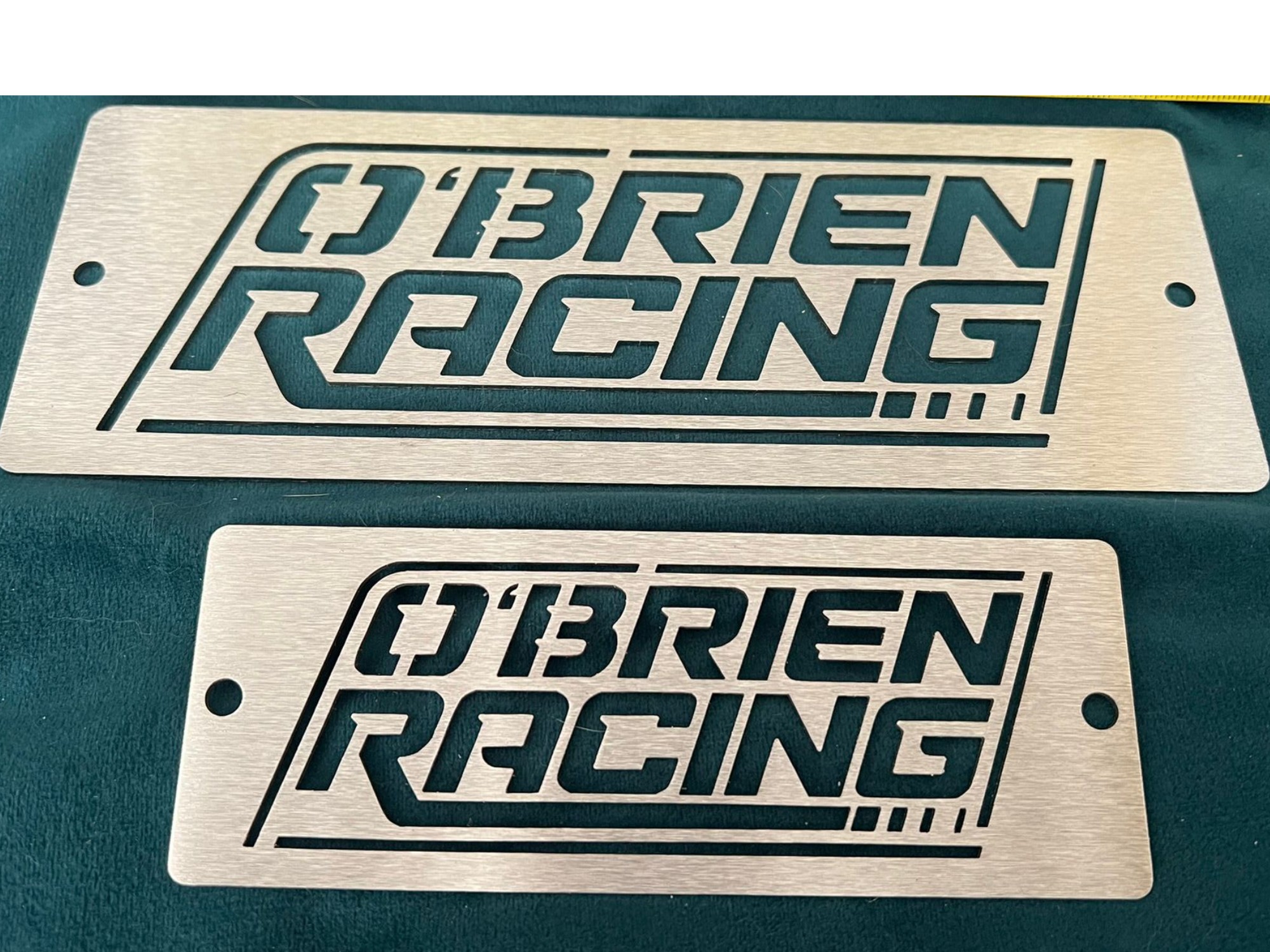 Two brushed metal O’Brien Racing branding plates with cut out lettering and mounting holes