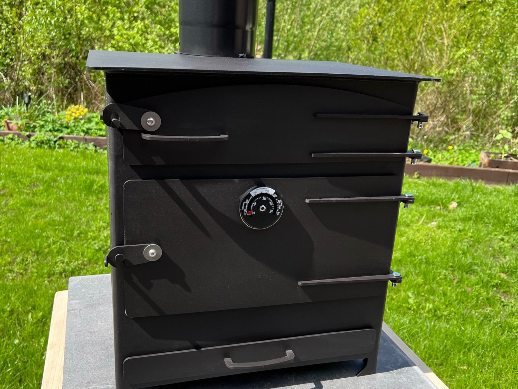 “Black outdoor wood‑fired oven with chimney, temperature gauge, and steel door in a garden setting