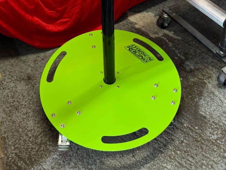 Green O’Brien Racing metal stand base with central pole and bolt‑mounted plate in a workshop