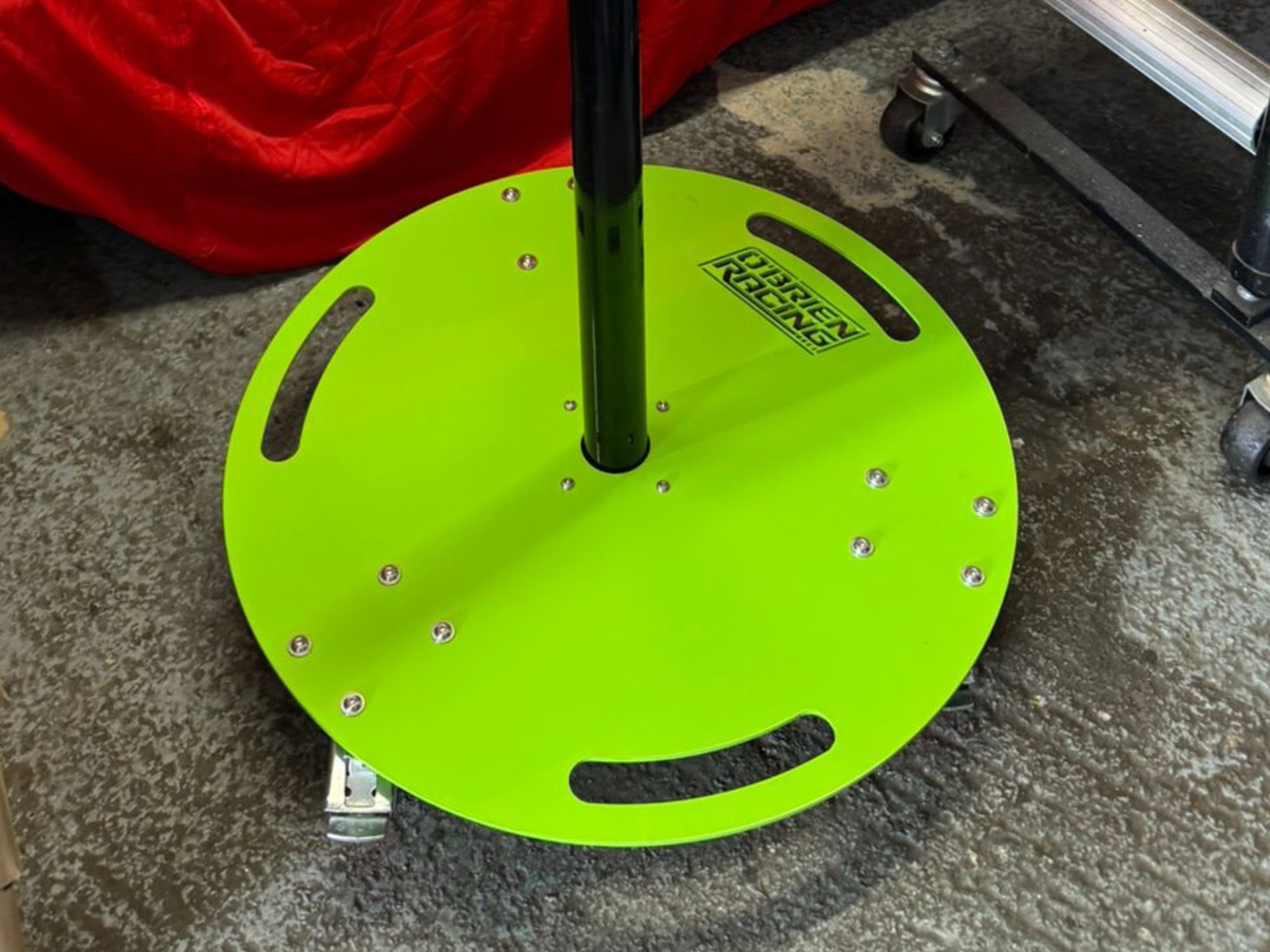 Green O’Brien Racing metal stand base with central pole and bolt‑mounted plate in a workshop