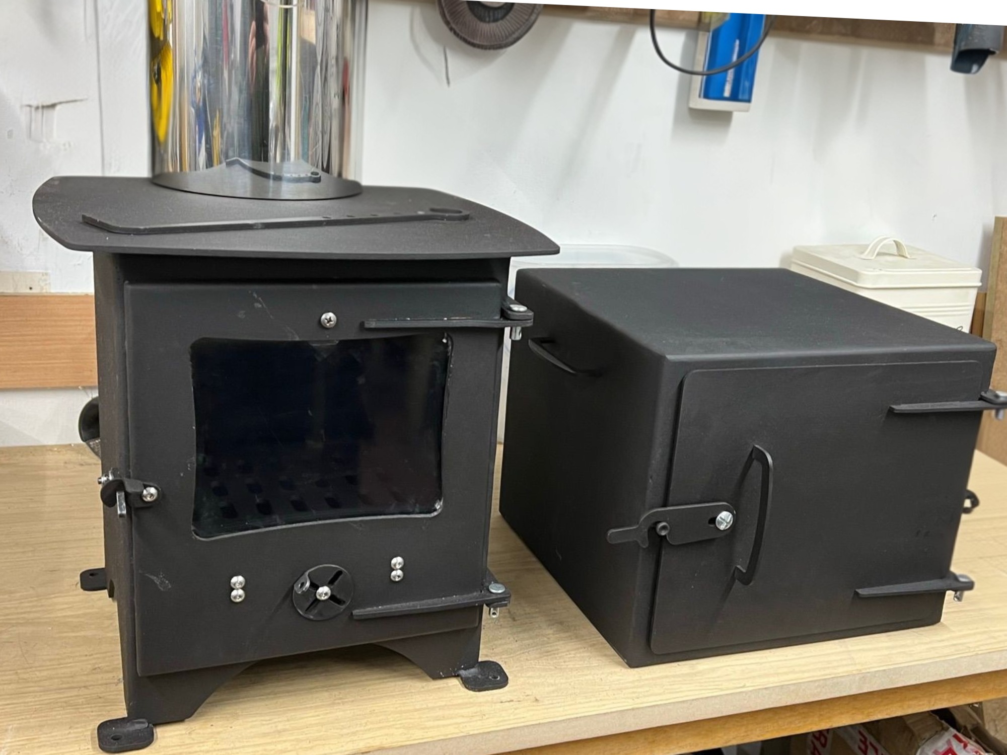 Compact black steel wood‑burning stove with glass door beside matching metal oven attachment in a workshop