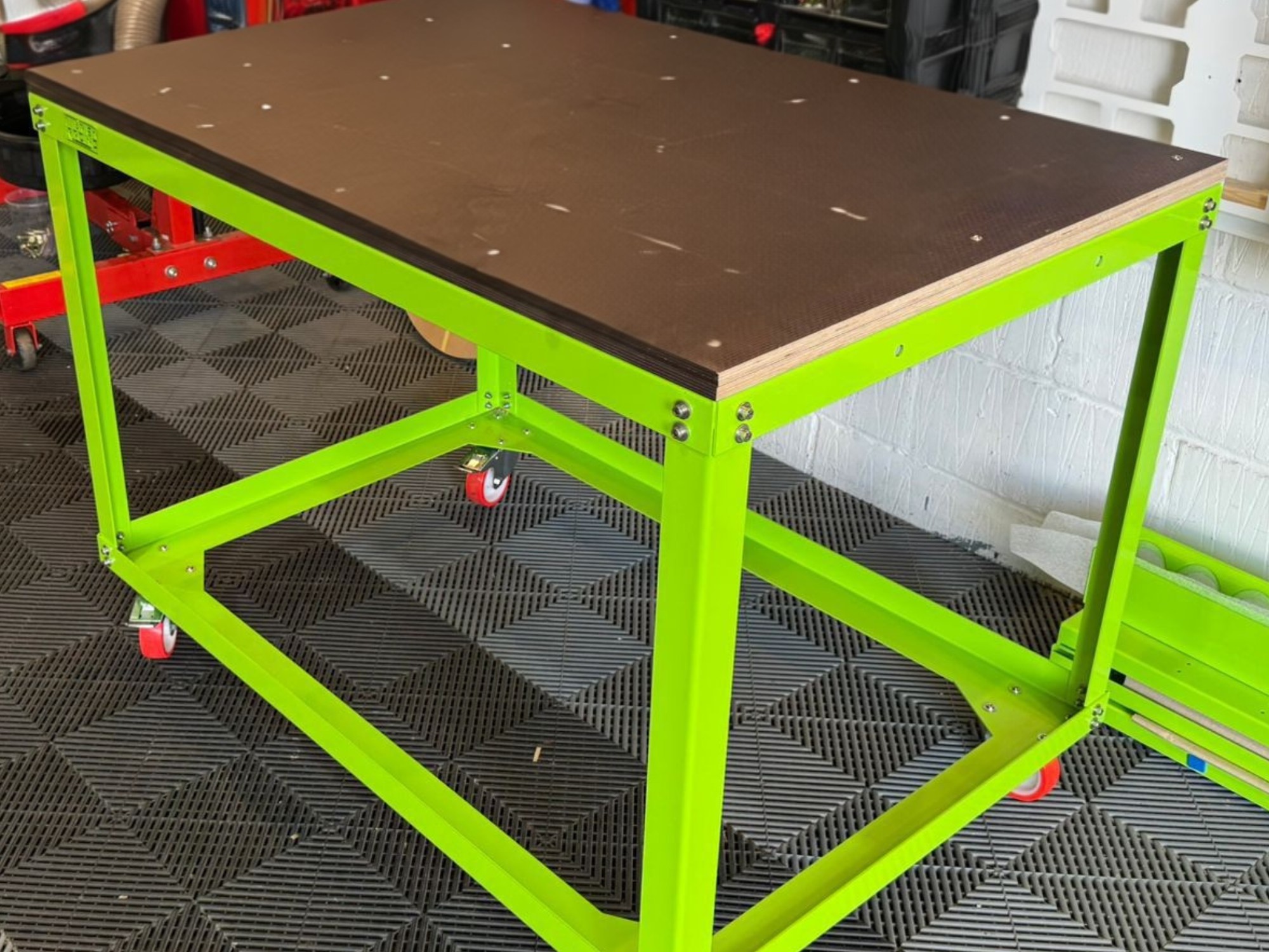 Green metal motorsport workbench with dark tabletop and red caster wheels in a workshop